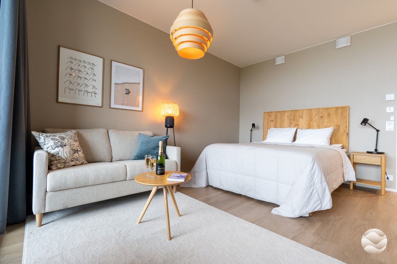 Hotellihuone, jossa on sänky, sohva, pieni pyöreä pöytä, jonka päällä on pullo ja laseja, lamppu sekä kaksi kehystettyä kuvaa seinällä.