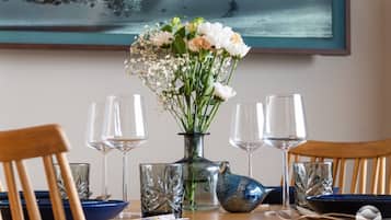 Een eettafel met een bloemenvaas, vier wijnglazen en een blauwe keramieken kruik.