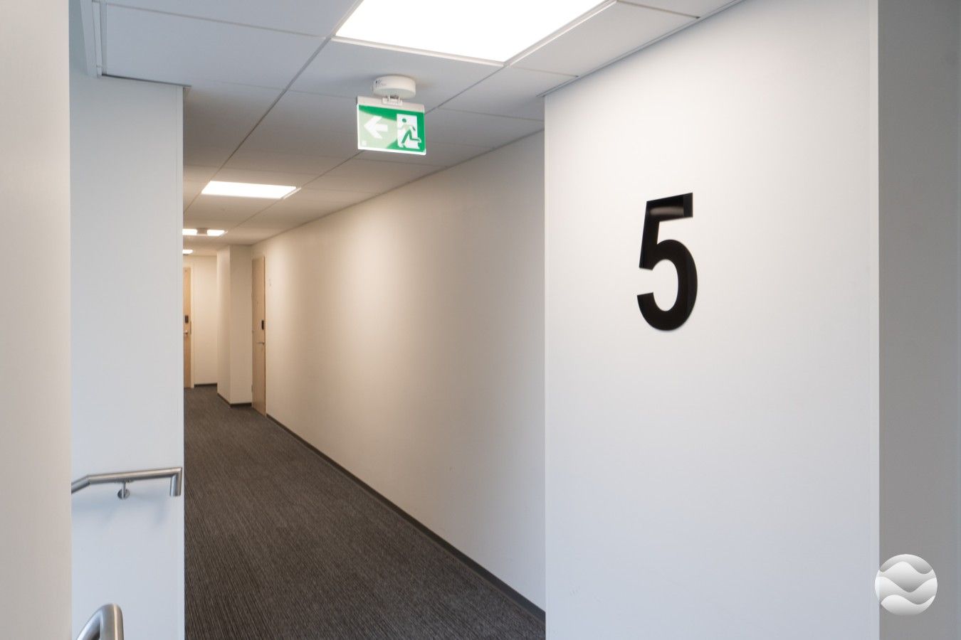 Un couloir avec un panneau portant le numéro 5 sur le mur, une porte au fond et un panneau indiquant la sortie de secours au plafond.