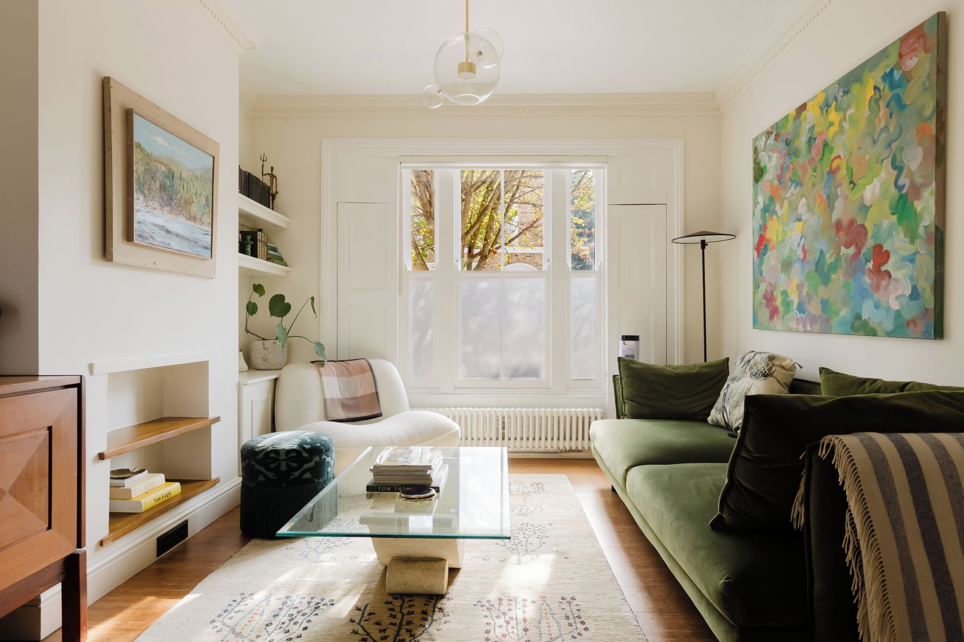 A living room with a glass coffee table, a green sofa, a white armchair, a bookshelf, and a large abstract painting.