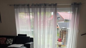 Un salon avec un canapé noir, un tapis rouge et une vue sur un espace extérieur doté d’un toit rouge, visible à travers des rideaux transparents.