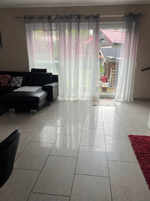 Ein Wohnzimmer mit einem schwarzen Sofa, einem roten Teppich und Blick durch transparente Vorhänge auf einen Außenbereich mit einem roten Dach.