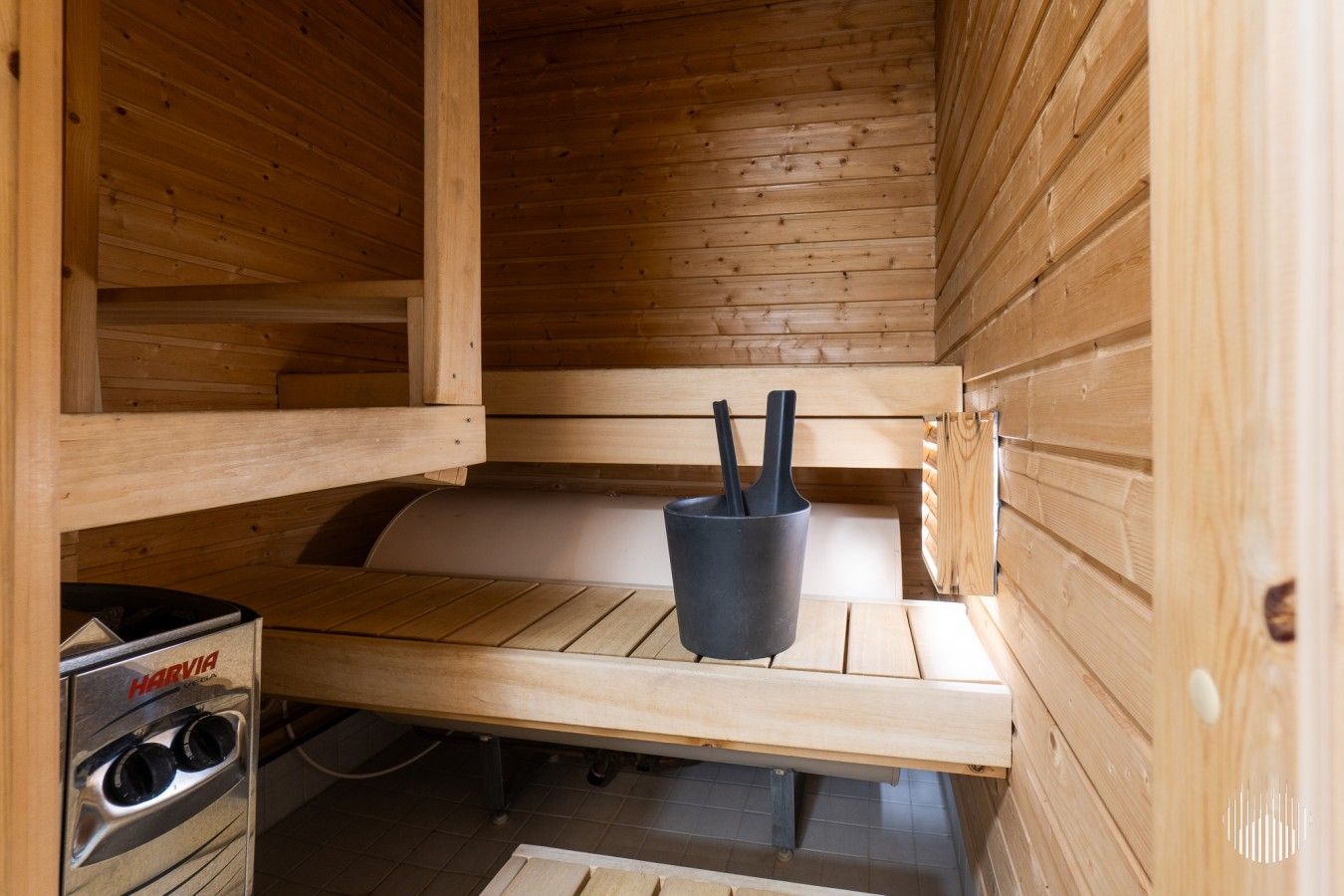 A sauna room with wooden walls and a bench. There is a sauna heater on the left and a black container with two handles on the bench.