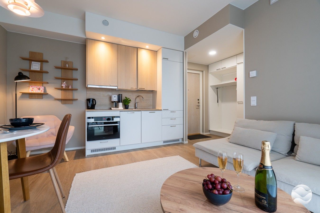 Een moderne keuken met witte kasten, een inbouwoven en een koffiezetapparaat. Er is een eethoek met een tafel en stoelen, en een woonkamer met een bank en een fles champagne.