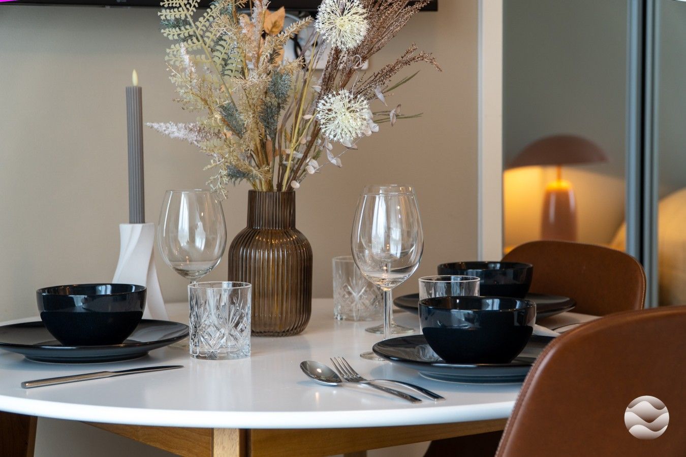 Une table à manger dressée avec des bols noirs, des verres transparents et un vase contenant des fleurs séchées.