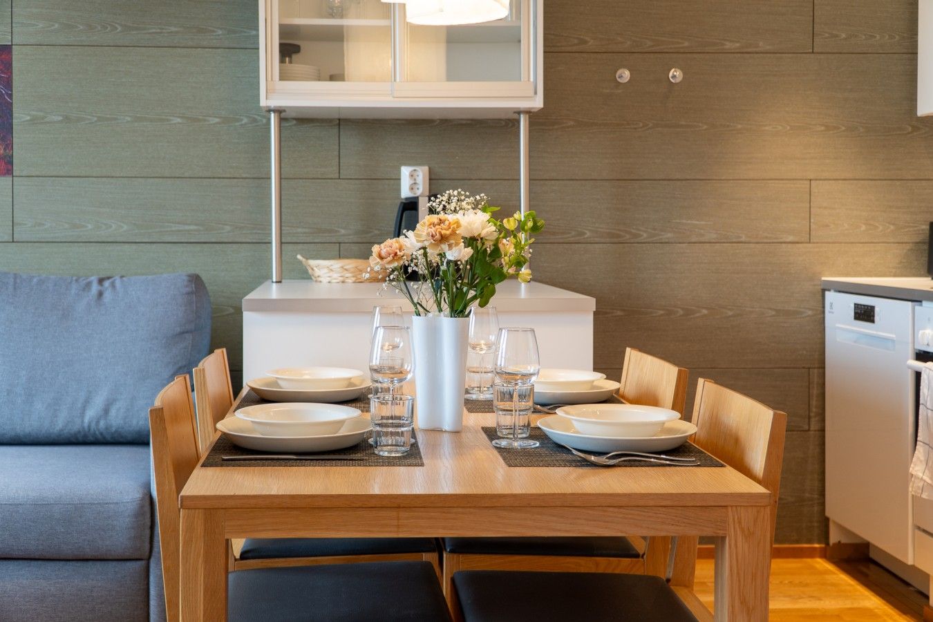 A dining area with a wooden table set for four, surrounded by chairs. A white vase with flowers is on the table, and there is a small side table with a basket.