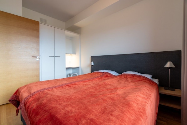 A neatly made bed with a red blanket, a bedside lamp, and a wardrobe with a mirror.