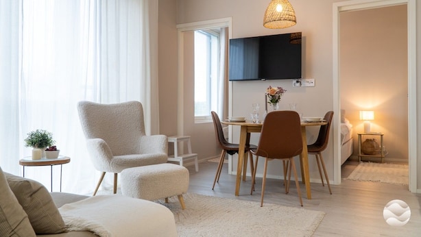 Una sala moderna con sofá, sillón, reposapiés, mesa de comedor con sillas, televisor montado en la pared y una lámpara sobre una mesita auxiliar.