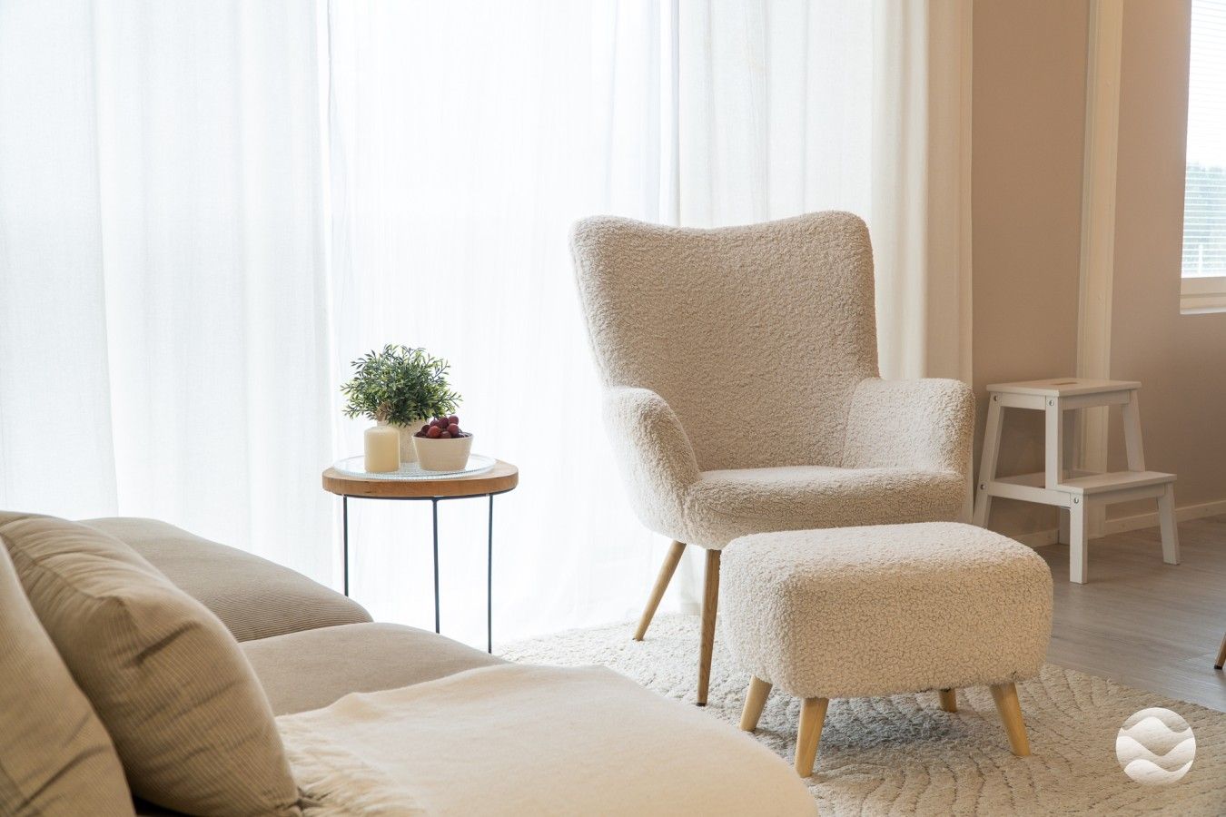 Un salon comprenant un canapé, un fauteuil moelleux, une petite table d’appoint avec une plante et un tabouret en bois.