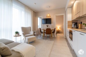 Una sala moderna con sofá, sillón, comedor con mesa y sillas, y cocina con armarios y electrodomésticos.
