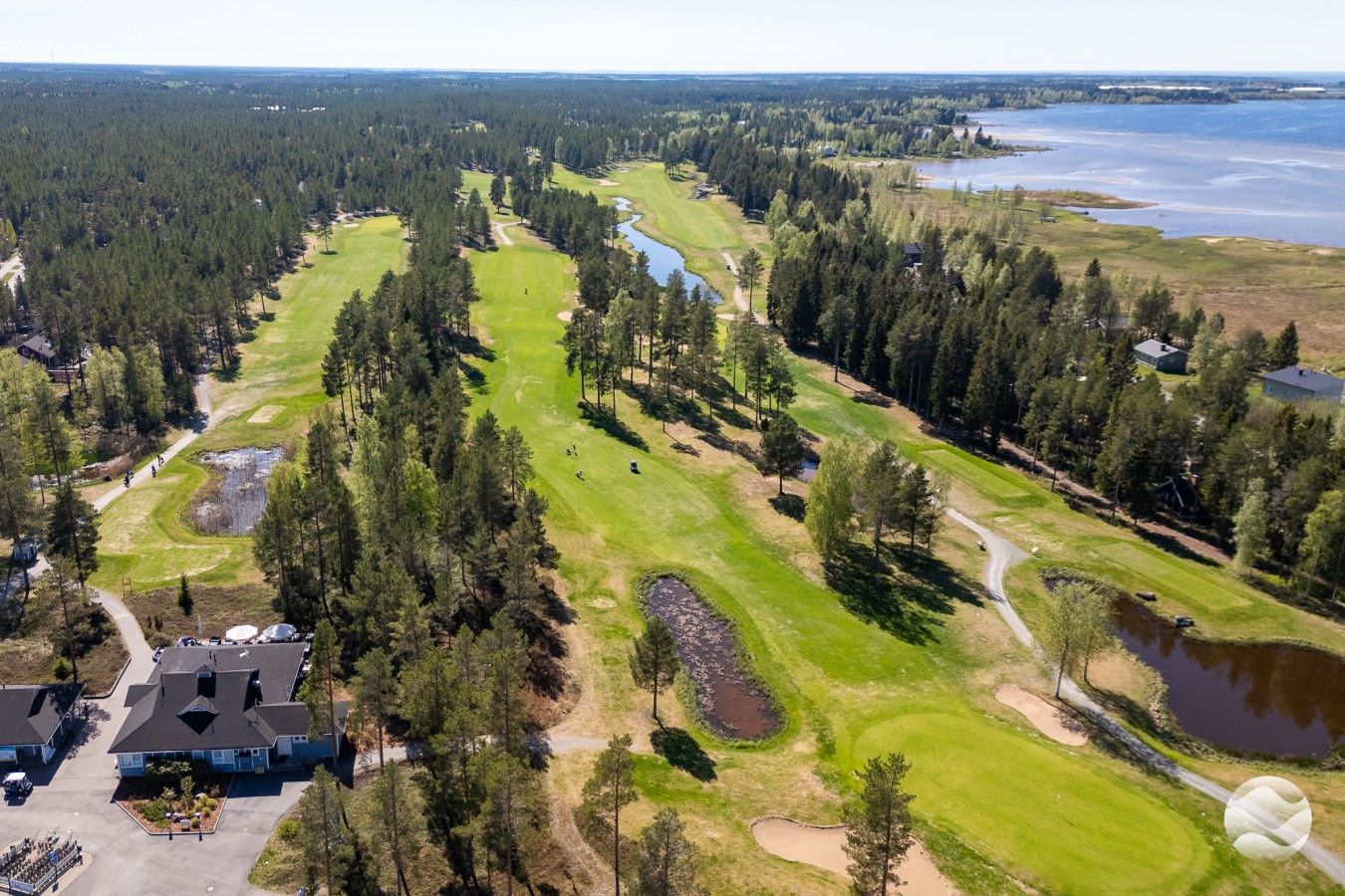 Een golfbaan met een vijver, omgeven door dicht bos en enkele gebouwen.