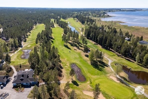 A golf course with a pond, surrounded by dense forest and a few buildings.