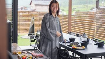 Una persona de pie junto a una parrilla con comida, una mesa con platos, y una vista de una cerca de madera y edificios al fondo.