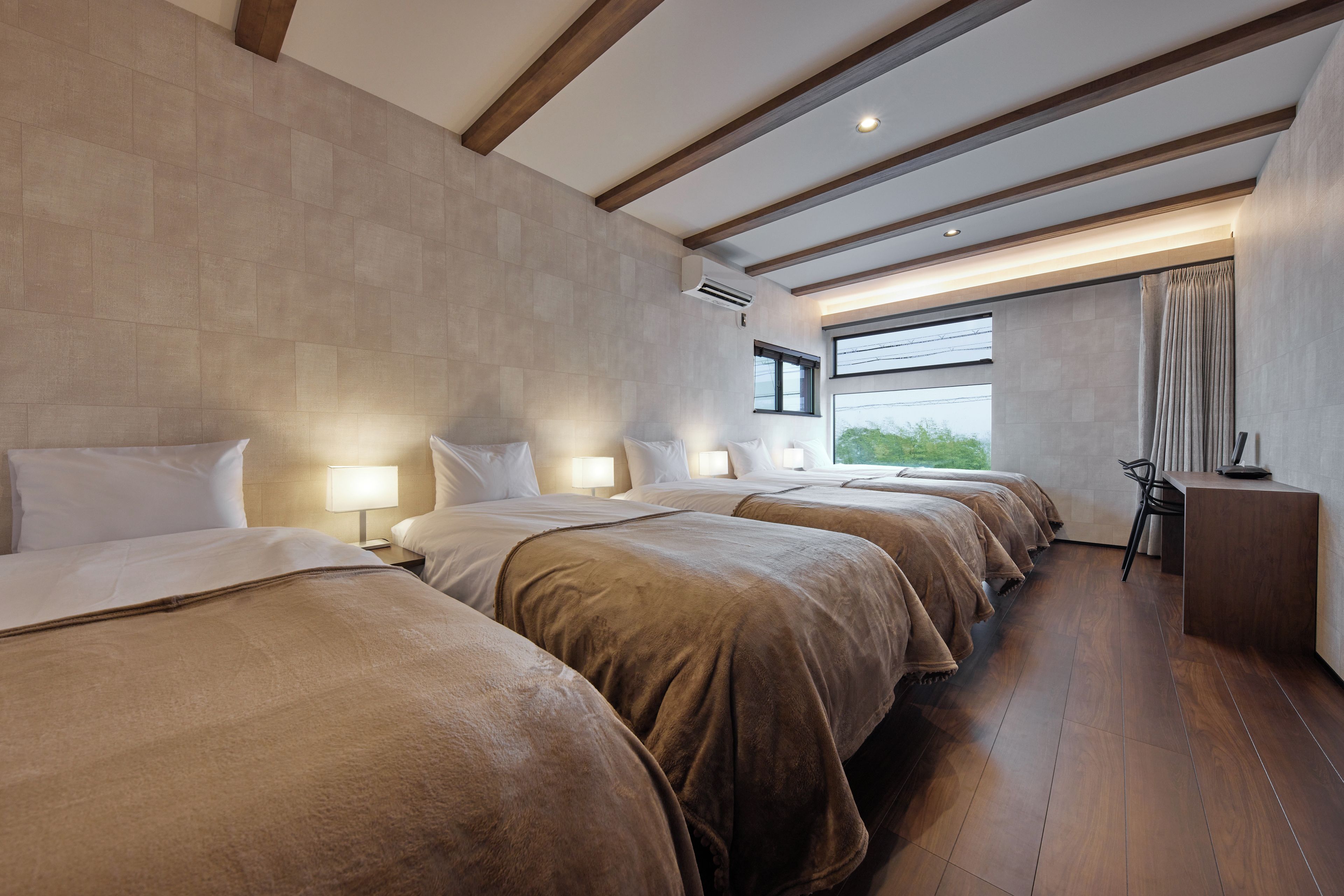 A hotel room with four beds aligned in a row, a large window with curtains, and a wooden floor.
