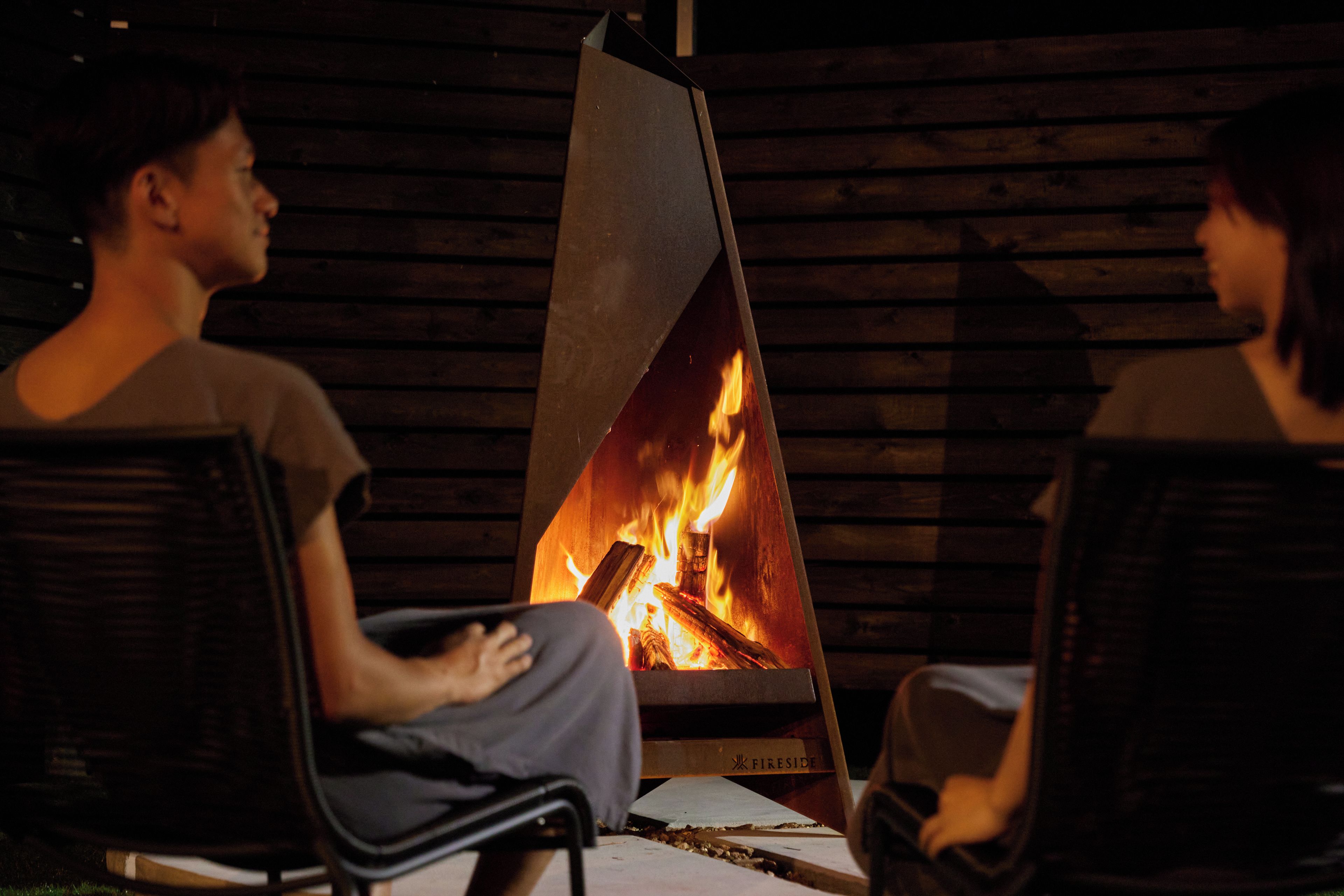 Dos personas sentadas en sillas frente a una chimenea de leña.
