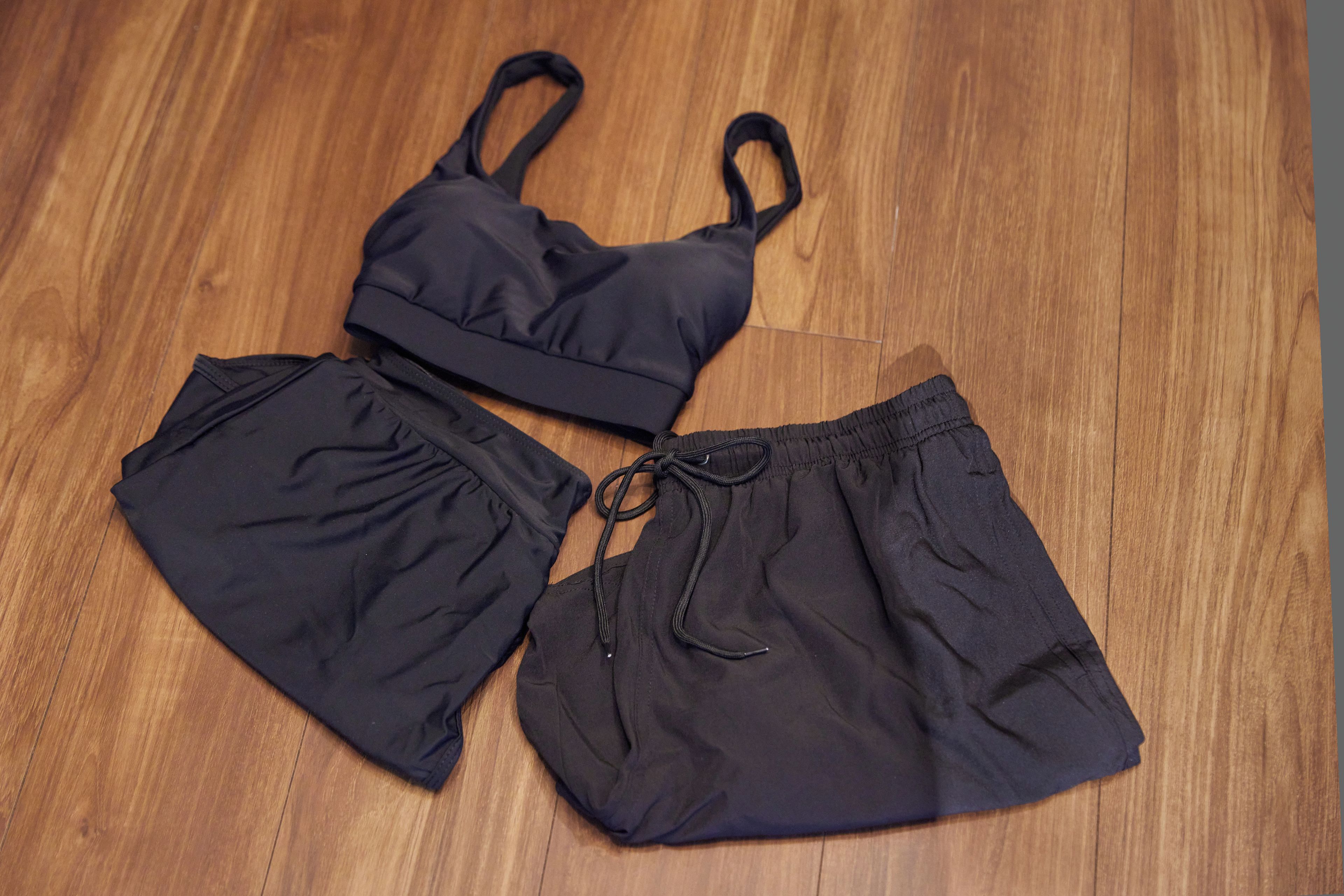 A pair of black shorts and a black bikini top laid out on a wooden floor.