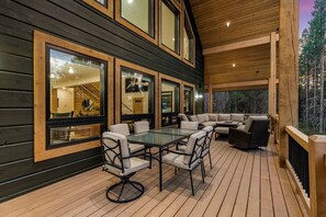 Una terraza de madera con una mesa y sillas de vidrio, un sofá y ventanas amplias.
