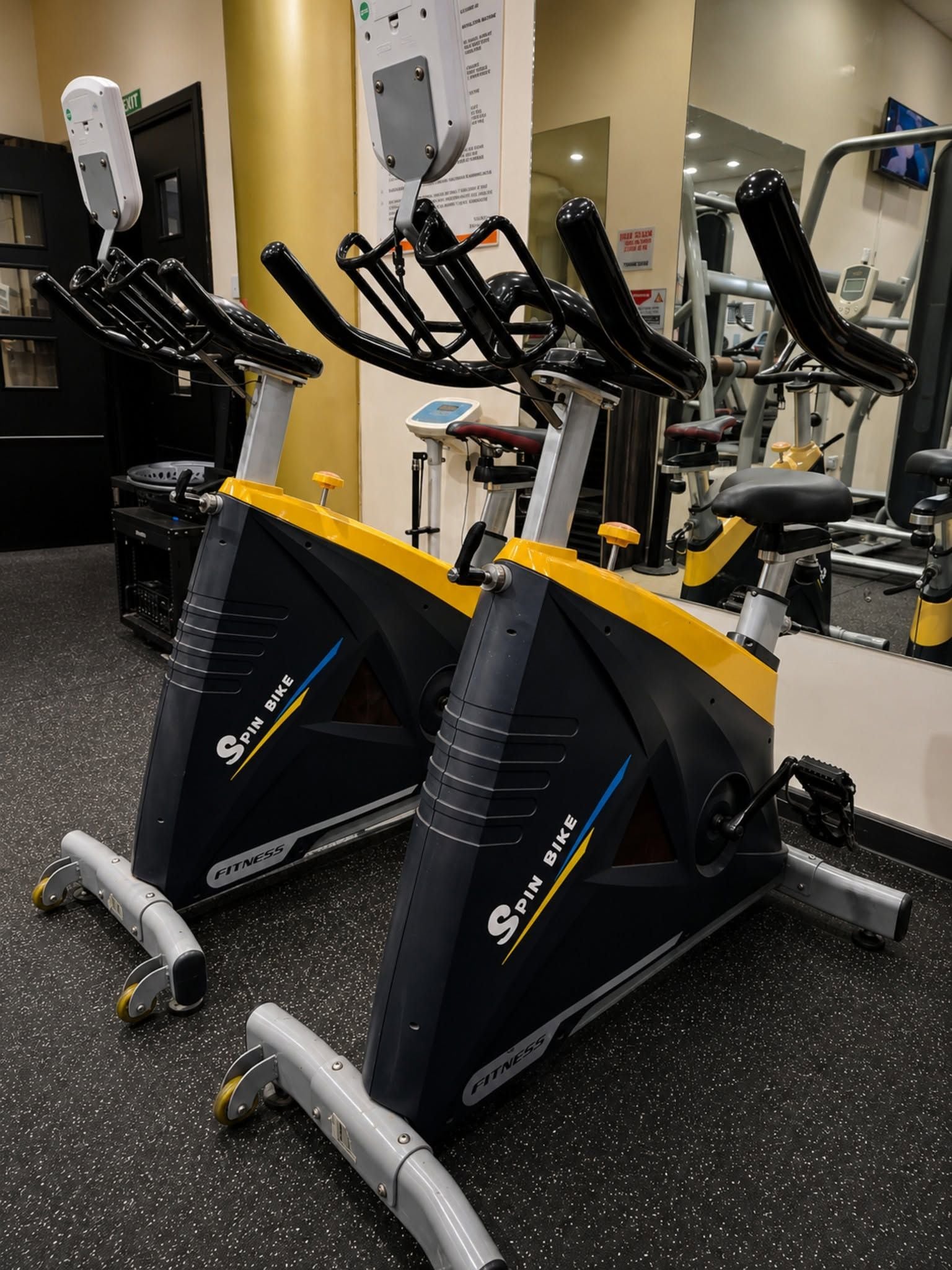 Dos bicicletas estáticas en un gimnasio con equipo de ejercicio al fondo.