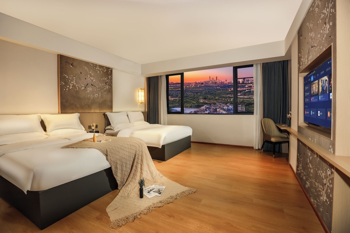 A hotel room with two beds, a large window with a city view, a flat-screen TV, and a wall art piece.