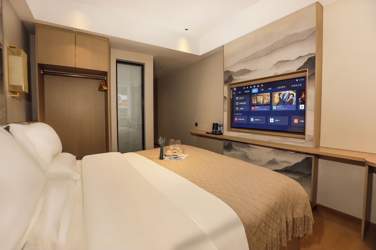 A hotel room with a bed, a TV mounted on the wall, a small table with a vase and glasses, and a door.