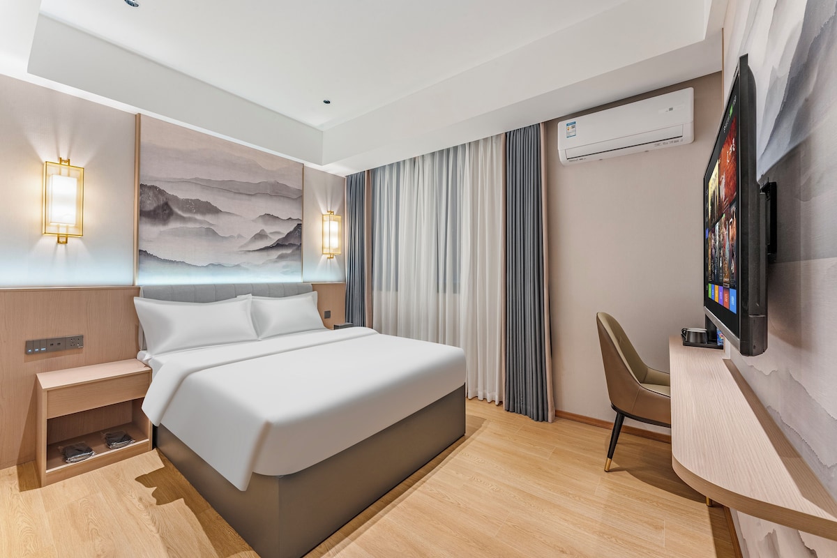 A modern hotel room with a bed, bedside tables, a desk with a chair, a mounted TV, and a large window with curtains.