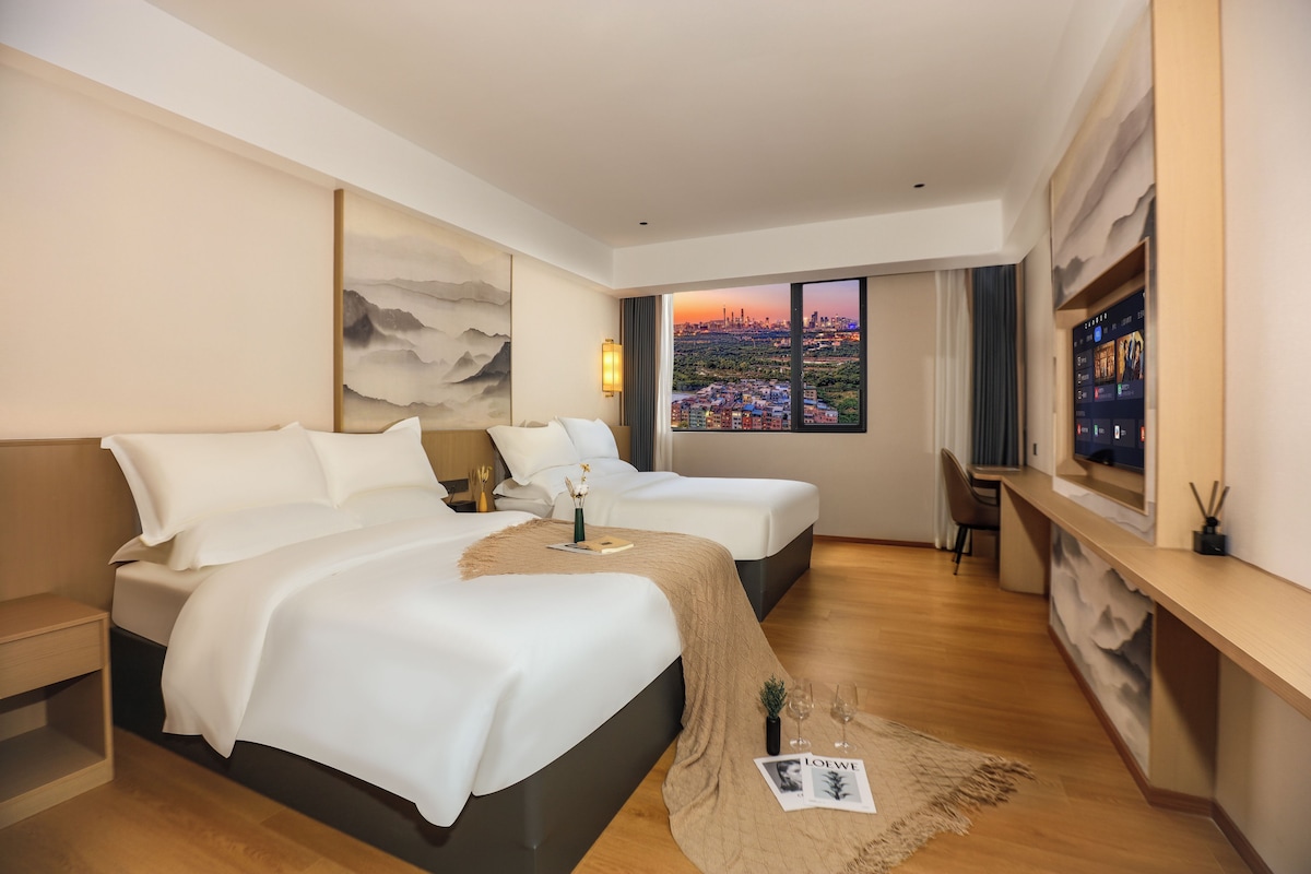 A hotel room with two beds, a desk, a large window with a city view, and a painting on the wall.