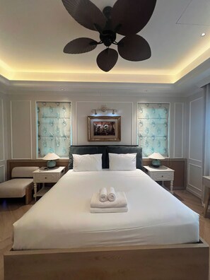 Una habitación de hotel con una cama grande, dos mesitas de noche con lámparas, un ventilador de techo y un cuadro enmarcado sobre la cama.