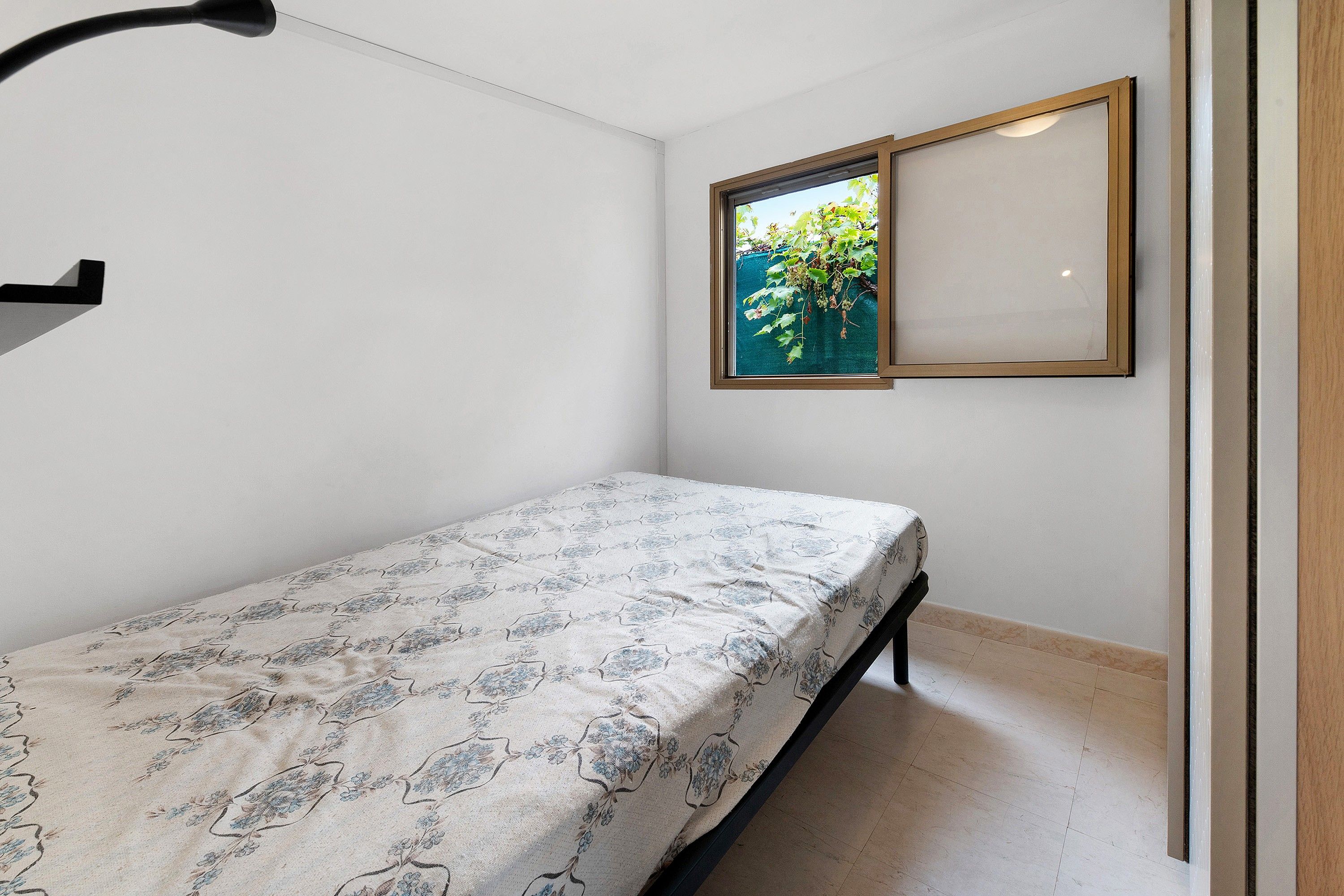 Une petite chambre avec un lit simple, une couvre-lit à motifs, un cadre photo accroché au mur et une fenêtre donnant sur un paysage verdoyant.