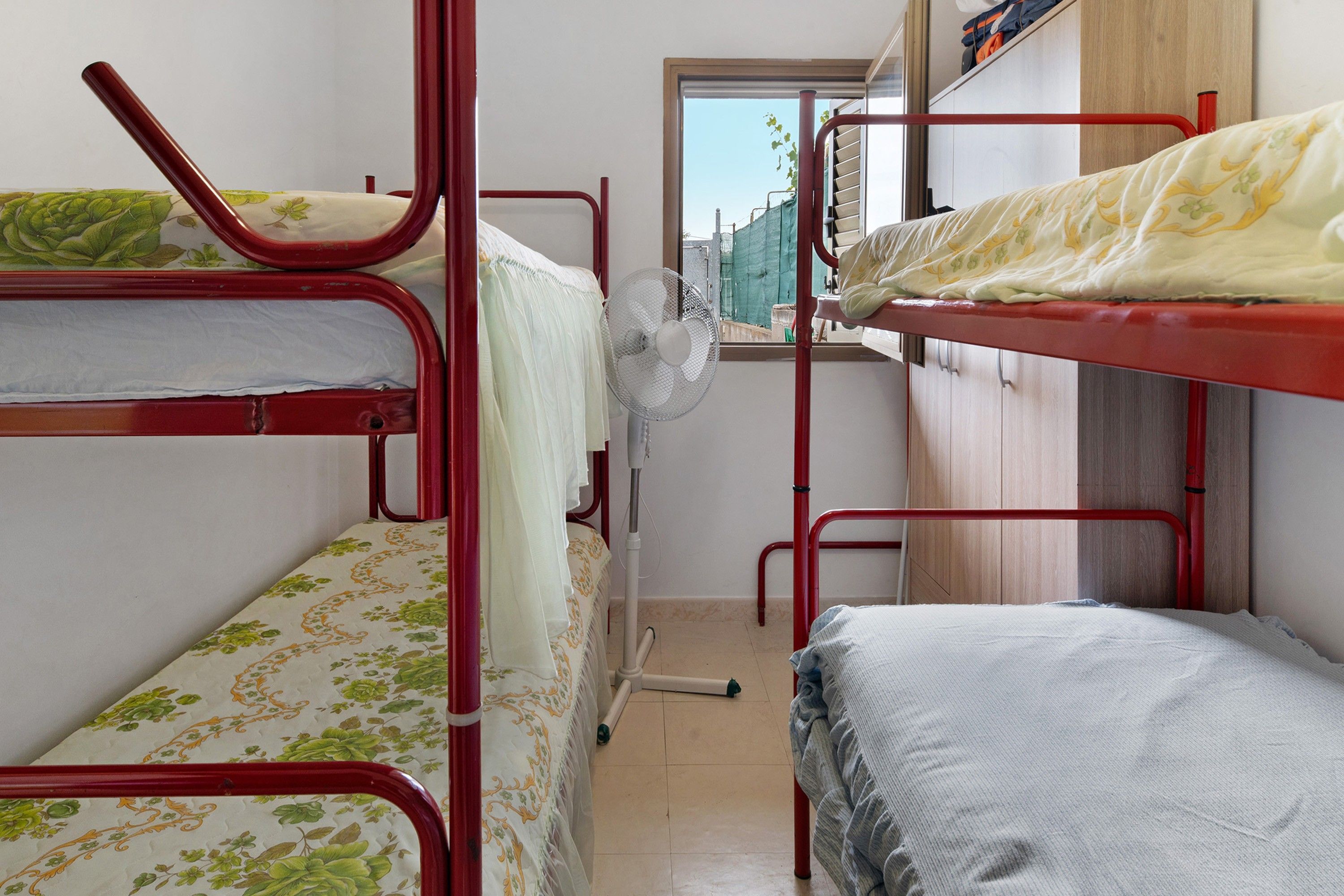 A dormitory room with bunk beds, a window, a fan, and a wooden wardrobe.
