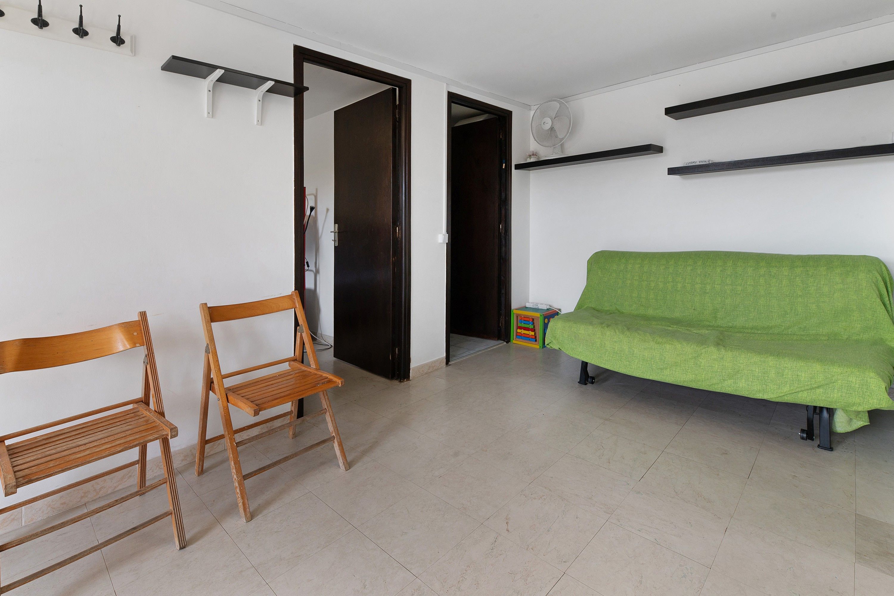Une pièce avec une chaise en bois, un canapé vert, une porte et un ventilateur fixé au mur.