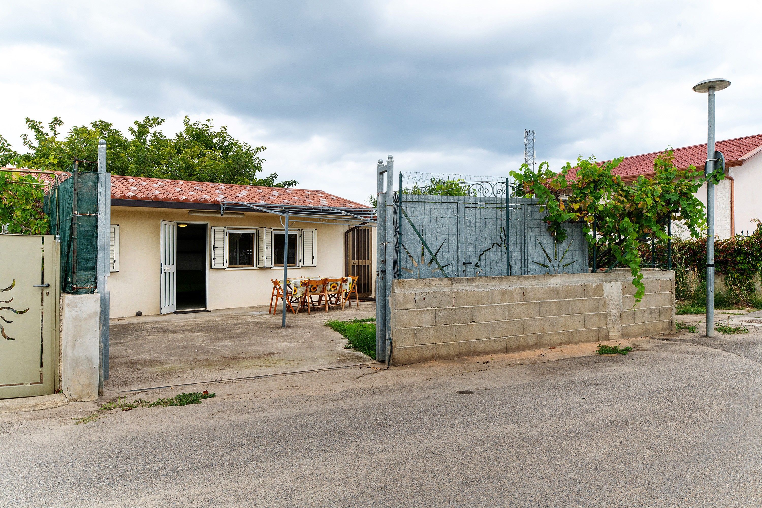 Une maison avec un toit recouvert de tuiles rouges, une entrée fermée par un portail et une table avec des chaises à l’extérieur.