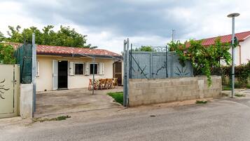 Une maison avec un toit recouvert de tuiles rouges, une entrée fermée par un portail et une table avec des chaises à l’extérieur.
