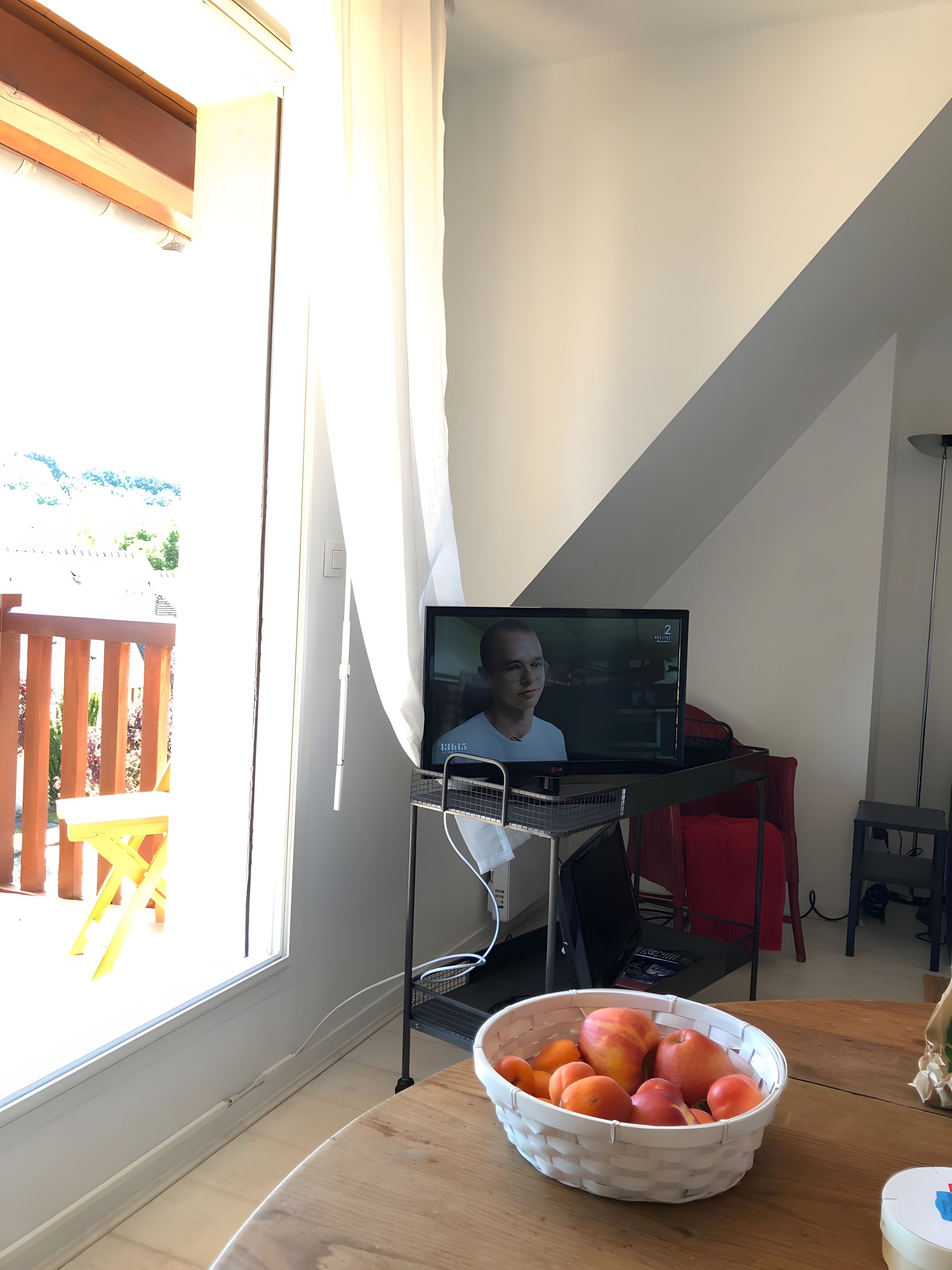 Una habitación con un ventanal grande, una computadora en un escritorio, una silla roja, una mesa de madera con una cesta de fruta, y vista a una terraza de madera.