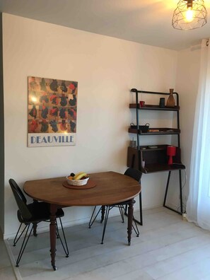 A dining area with a wooden table, black chairs, a fruit bowl, a wall-mounted shelf with various items, and a wall art piece labeled 'DEAUVILLE'. - Holiday Home '2 Pièces À Deauville Avec Piscine' with Wi-Fi (Deauville)