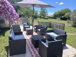 Een terras buiten met rieten meubels, een kamerkleed met dessin en een grote parasol.