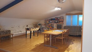 Een gezellig zolderkamer met een houten tafel, stoelen, een tafelvoetbaltafel en een verzameling speelgoed en spellen op planken.