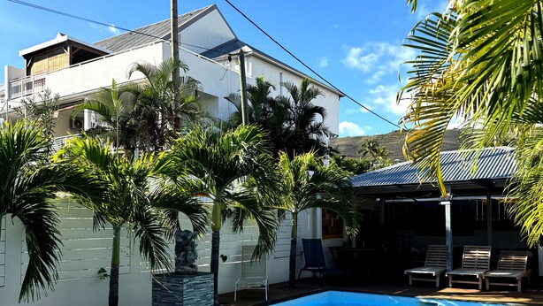 A swimming pool with a wooden deck, surrounded by palm trees and a white building with a sloped roof. - Côté Plage (Saint-Paul)