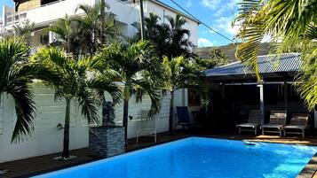 Una piscina con cubierta de madera, rodeada de palmeras y un edificio blanco con techo inclinado.