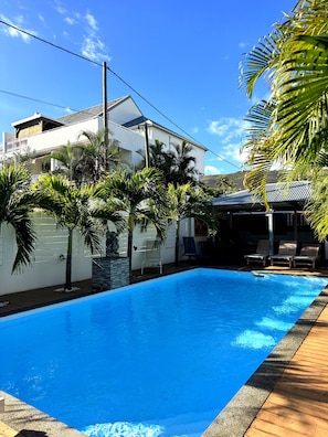 A swimming pool with a wooden deck, surrounded by palm trees and a white building with a sloped roof.
