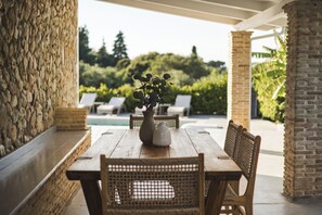 Una mesa de madera con sillas, un florero con una planta y vista a una piscina y follaje.