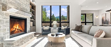 A modern living room with a stone fireplace, a large window with a view of trees, a sofa, and a coffee table.