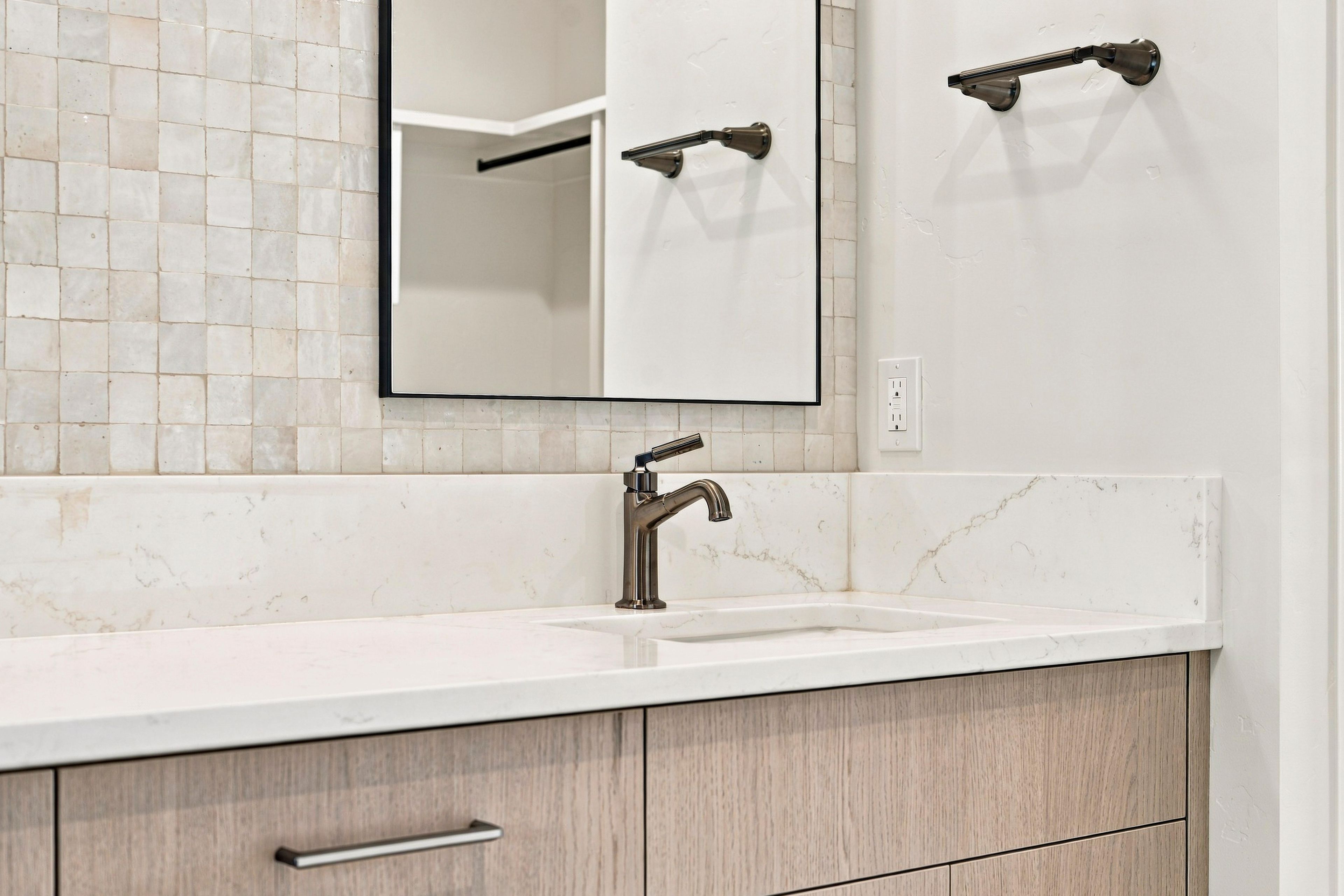 Un baño con salpicadero de azulejos blancos, un espejo, un lavamanos con grifo moderno y muebles de madera.