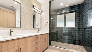 Ein modernes Badezimmer mit einem Doppelwaschtisch, einem großen Spiegel und einer begehbaren Dusche mit dunklen Fliesen.