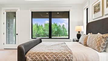A bedroom with a large window, a bed with patterned bedding, and a nightstand with a lamp.