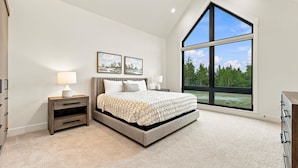 A bedroom with a large bed, two bedside tables, a nightstand, a window with a view of trees, and two wall-mounted paintings.