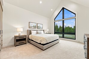 Ein Schlafzimmer mit einem großen Bett, zwei Nachttischen, einem Fenster mit Blick auf Bäume und zwei an der Wand befestigten Bildern.
