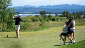 A golf course with a player preparing to hit a ball, a caddie, and a woman walking beside them.