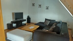 Una sala de estar con un televisor de pantalla plana, un sofá, una mesa de centro y un pequeño reposapiés blanco.