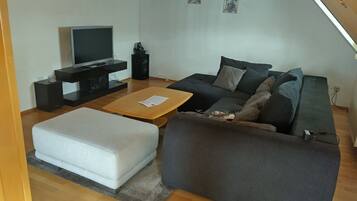 Een woonkamer met een flatscreen televisie, een bank, een salontafel en een klein, wit poufje.