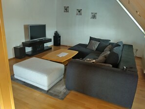 Uma sala de estar com televisão de tela plana, sofá, mesa de centro e um banquinho branco.